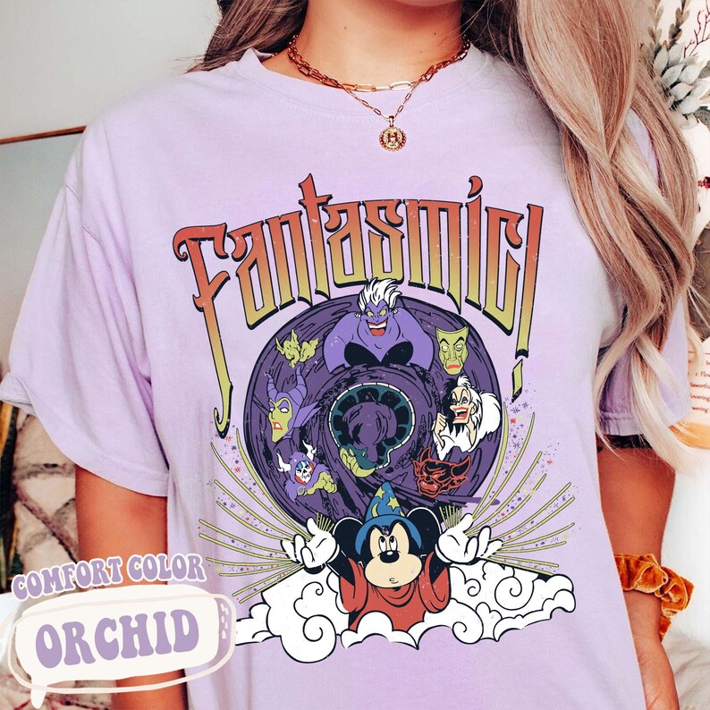 May include: Orchid-colored t-shirt with a graphic featuring the word "Fantasmic!" in a retro font. The design includes cartoon characters, including Mickey Mouse in a sorcerer's outfit and various villains. The shirt is labeled "Comfort Color".