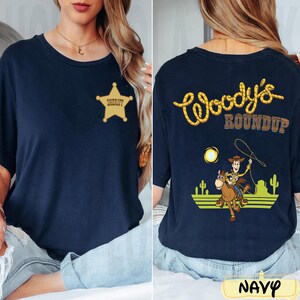 K&ouml;nnte beinhalten: Marineblaues T-Shirt mit goldenem Sheriff-Abzeichen auf der Vorderseite. Auf der R&uuml;ckseite steht "Woody's Roundup" in gelben Buchstaben im Seil-Stil, mit einem Cartoon von Woody auf einem Pferd, einer Sonne und Kakteen.