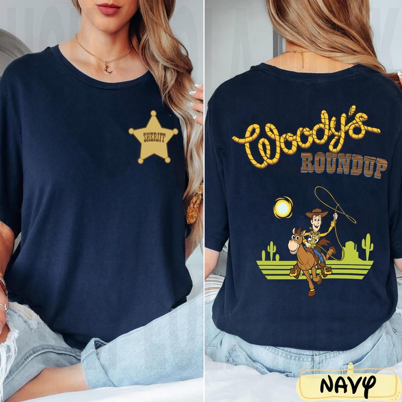K&ouml;nnte beinhalten: Marineblaues T-Shirt mit goldenem Sheriff-Abzeichen auf der Vorderseite. Auf der R&uuml;ckseite steht "Woody's Roundup" in gelben Buchstaben im Seil-Stil, mit einem Cartoon von Woody auf einem Pferd, einer Sonne und Kakteen.