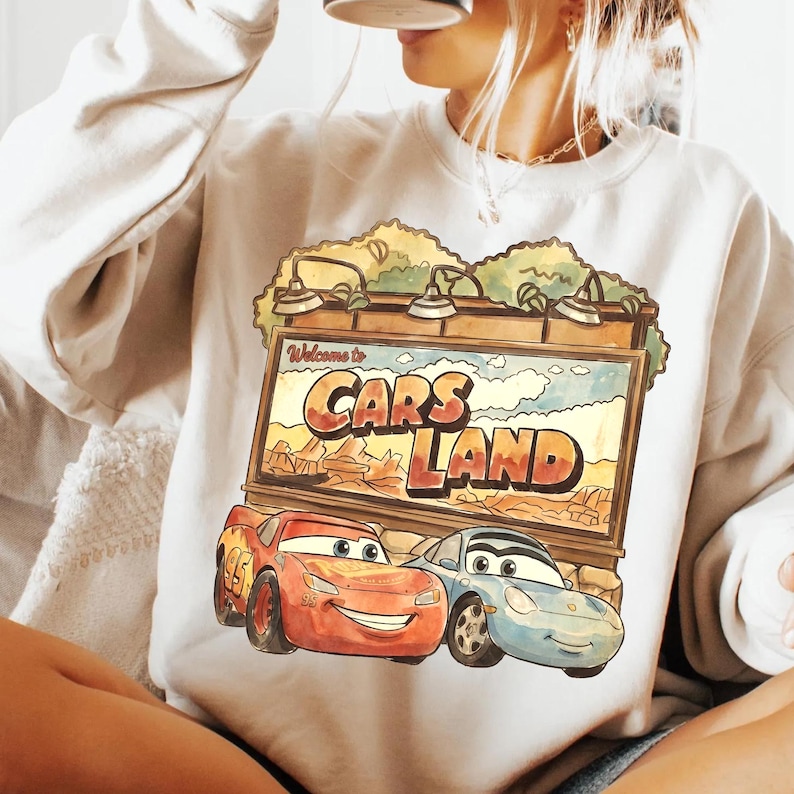 Op de afbeelding: Cr&egrave;mekleurige sweatshirt met een afbeelding van twee cartoonauto's voor een bord met de tekst "Welcome to Cars Land". Het bord staat tegen een woestijnlandschap. De auto's zijn rood en blauw.