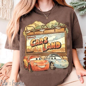 Op de afbeelding: Espresso-kleurig T-shirt met een afbeelding van het Cars Land-bord met Bliksem McQueen en Sally Carrera. Het bord leest "Welcome to Cars Land". Het shirt heeft een comfortabele pasvorm.