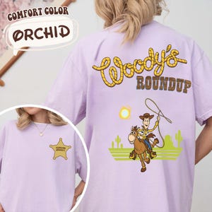 K&ouml;nnte beinhalten: Orchideenfarbenes T-Shirt mit einem Sheriff-Abzeichen auf der Vorderseite und einer Woody's Roundup-Grafik auf der R&uuml;ckseite. Die Grafik auf der R&uuml;ckseite zeigt einen Cowboy auf einem Pferd mit Lasso, Sonne und Kakteen. Das Shirt ist mit "Comfort Color" gekennzeichnet.