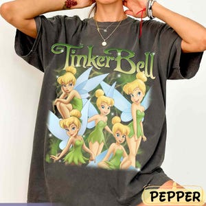 May include: Dark gray t-shirt featuring the text "Tinker Bell" in green with a light green and white design. The shirt has a graphic of Tinker Bell and other fairies in green dresses with blue wings.