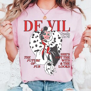May include: Pink t-shirt featuring a graphic of Cruella De Vil with the text "DE VIL" in red. The design includes the phrases "THE FUTURE OF FUR" and "SHE'S GOT THE WITS & THE LOOKS".