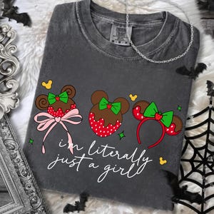 May include: Dark gray t-shirt with a graphic of three mouse-eared designs, each with a strawberry and bow. The text "i'm literally just a girl" is written in cursive. The shirt is a Custom Colors brand.