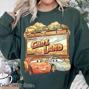Op de afbeelding: Bosse groen sweatshirt met een Cars Land-afbeelding. De afbeelding bevat de tekst "Welcome to Cars Land" boven illustraties van Bliksem McQueen en Sally Carrera. De sweatshirt is effen van kleur.