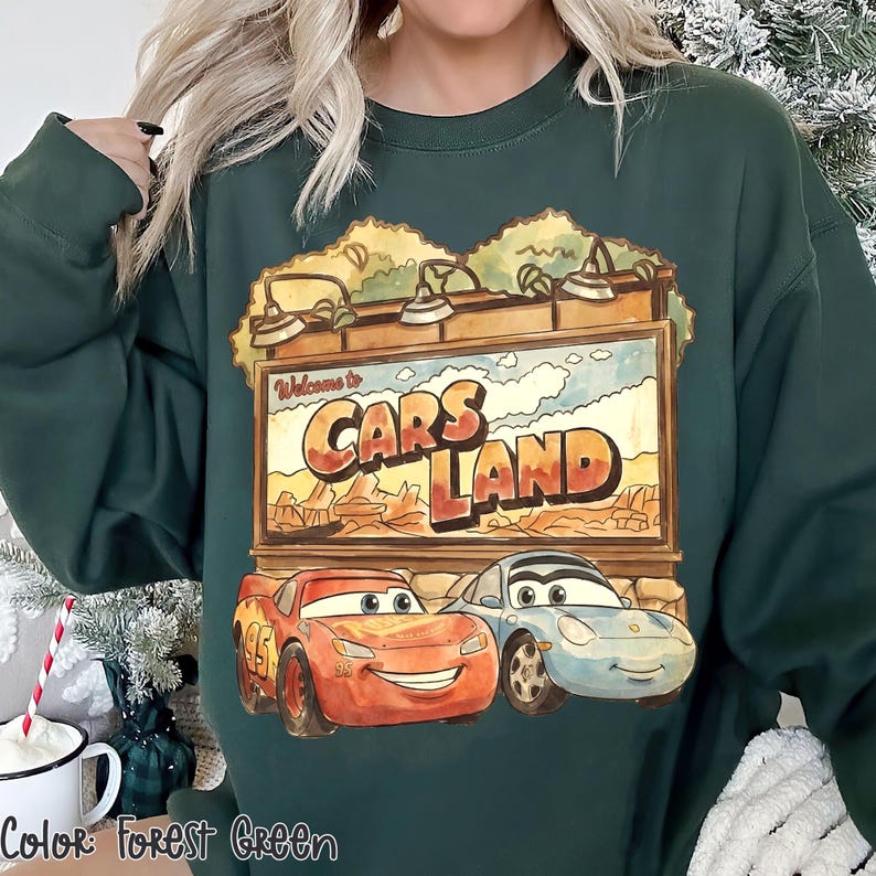 Op de afbeelding: Bosse groen sweatshirt met een Cars Land-afbeelding. De afbeelding bevat de tekst "Welcome to Cars Land" boven illustraties van Bliksem McQueen en Sally Carrera. De sweatshirt is effen van kleur.