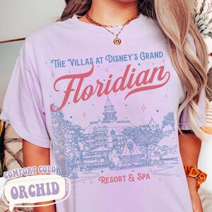 May include: Orchid-colored t-shirt with a graphic design featuring the text "The Villas at Disney's Grand Floridian Resort & Spa." The design includes an illustration of the resort in blue and red lettering. The shirt also has the words "Comfort Color" and "Orchid" on it.