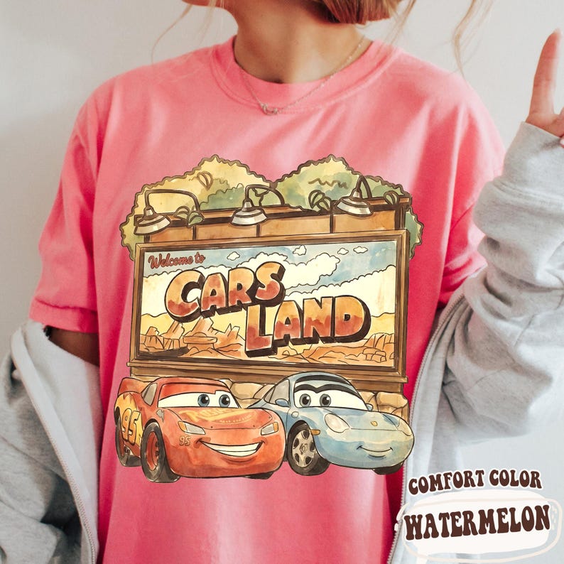 Op de afbeelding: Roze T-shirt met een Cars Land-ontwerp. De afbeelding bevat de tekst "Welcome to Cars Land" boven een afbeelding van Bliksem McQueen en Sally Carrera. Het shirt is een Comfort Color in Watermelon.