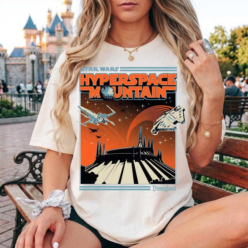 May include: Cream-colored t-shirt featuring a Star Wars "Hyperspace Mountain" graphic. The design includes spaceships, a mountain silhouette, and the Disneyland logo. The text is in orange and blue.