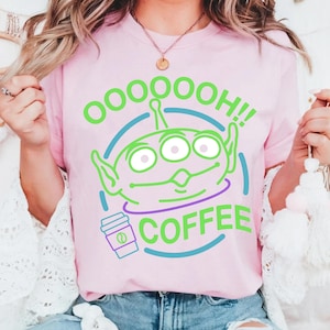 Puede incluir: Camiseta rosa claro con un gr&aacute;fico verde y azul de un extraterrestre de tres ojos con las palabras "OOOOOOH!! COFFEE". Tambi&eacute;n se incluye un gr&aacute;fico de taza de caf&eacute;. La camiseta es de cuello redondo.