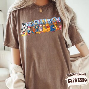 Puede incluir: Camiseta marr&oacute;n con un gr&aacute;fico de personajes de Disney y la palabra "Disneyland" en letras coloridas. La camiseta est&aacute; etiquetada como "Comfort Color Espresso" en una fuente estilizada.