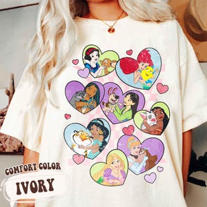 Puede incluir: Camiseta de color marfil con ilustraciones en forma de corazón de personajes animados. Los corazones contienen imágenes de princesas y sus compañeros animales. Las palabras "COMFORT COLOR" e "IVORY" están impresas en la parte inferior izquierda.