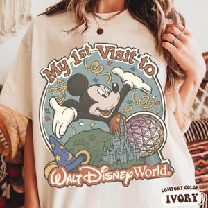 May include: Ivory-colored t-shirt featuring a graphic of Mickey Mouse with the text "My 1st Visit to Walt Disney World." The design includes a castle, a sorcerer's hat, and a geometric sphere. The shirt is labeled "Comfort Color" and "Ivory."