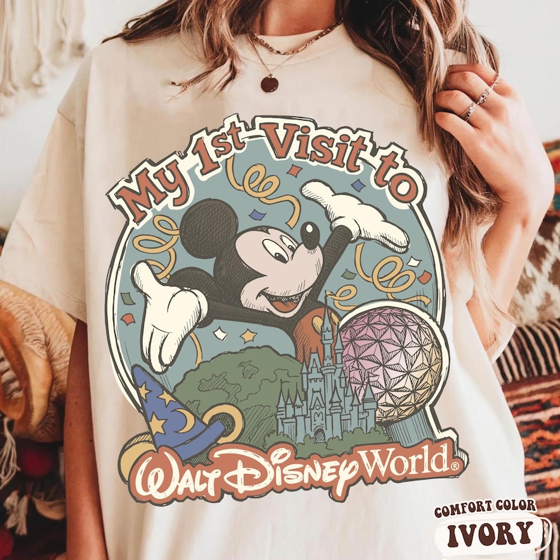 May include: Ivory-colored t-shirt featuring a graphic of Mickey Mouse with the text "My 1st Visit to Walt Disney World." The design includes a castle, a sorcerer's hat, and a geometric sphere. The shirt is labeled "Comfort Color" and "Ivory."