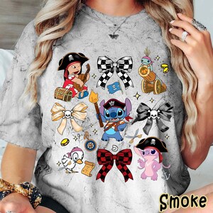 Może przedstawiać: Szara koszulka typu tie-dye z postaciami pirat&oacute;w z kresk&oacute;wek i kokardkami. Projekt zawiera pirackie dziecko grające na gitarze, Stitcha z pochodnią i r&oacute;żowego pirata-kosmitę. Słowo "Smoke" jest wydrukowane na dole.