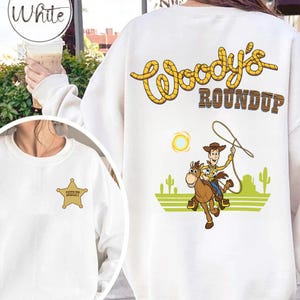 K&ouml;nnte beinhalten: Wei&szlig;es Sweatshirt mit Sheriff-Abzeichen und einer Grafik von Woody aus Toy Story auf einem Pferd, mit dem Text "Woody's Roundup". Das Design umfasst eine Sonne, Kakteen und einen Lasso.