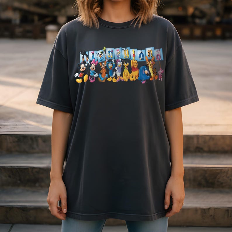 Puede incluir: Camiseta gris oscuro con un estampado colorido de personajes de Disney, incluyendo Mickey Mouse, Donald Duck y Winnie the Pooh. La palabra "Disneyland" est&aacute; impresa encima de los personajes. La camiseta tiene mangas cortas y un corte holgado.