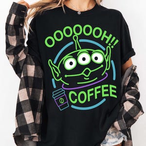 Puede incluir: Camiseta negra con un gr&aacute;fico de alien&iacute;gena verde y las palabras "OOOOOOH!! COFFEE" en verde y blanco. El alien&iacute;gena tiene tres ojos y orejas grandes. Tambi&eacute;n aparece una taza de caf&eacute; morada.