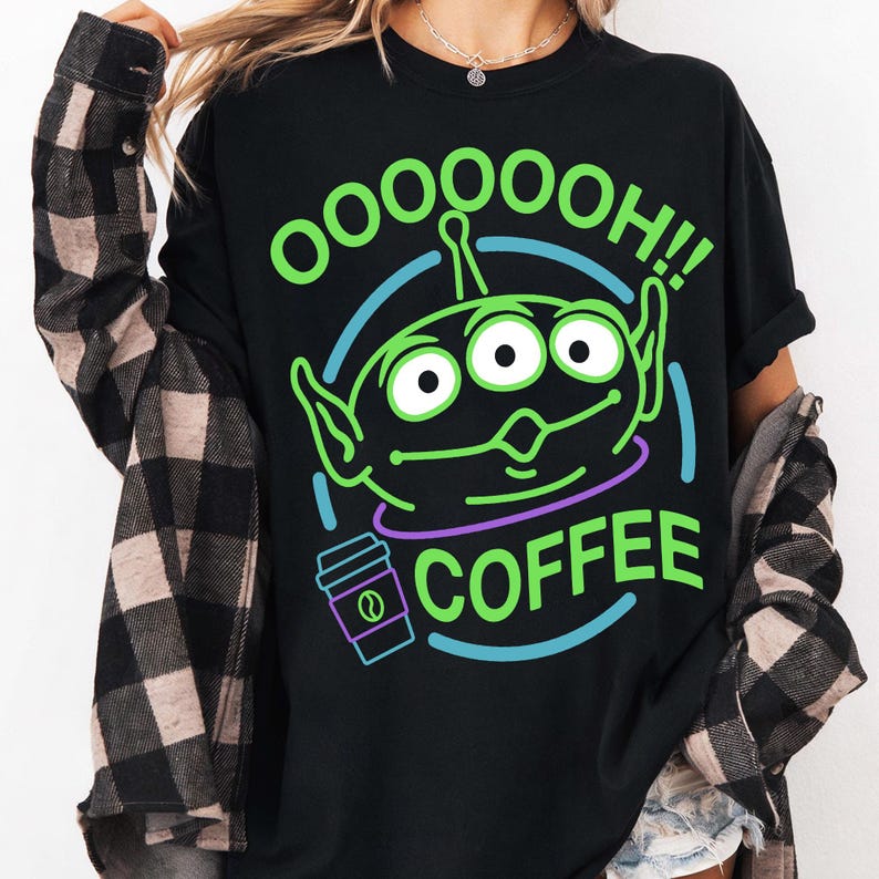 Puede incluir: Camiseta negra con un gr&aacute;fico de alien&iacute;gena verde y las palabras "OOOOOOH!! COFFEE" en verde y blanco. El alien&iacute;gena tiene tres ojos y orejas grandes. Tambi&eacute;n aparece una taza de caf&eacute; morada.