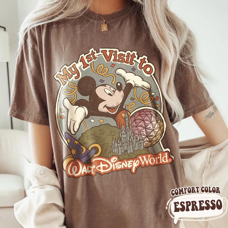May include: Espresso-colored t-shirt featuring a vintage-style graphic of Mickey Mouse celebrating his first visit to Walt Disney World. The design includes a castle, a sorcerer's hat, and the text "My 1st Visit to Walt Disney World."