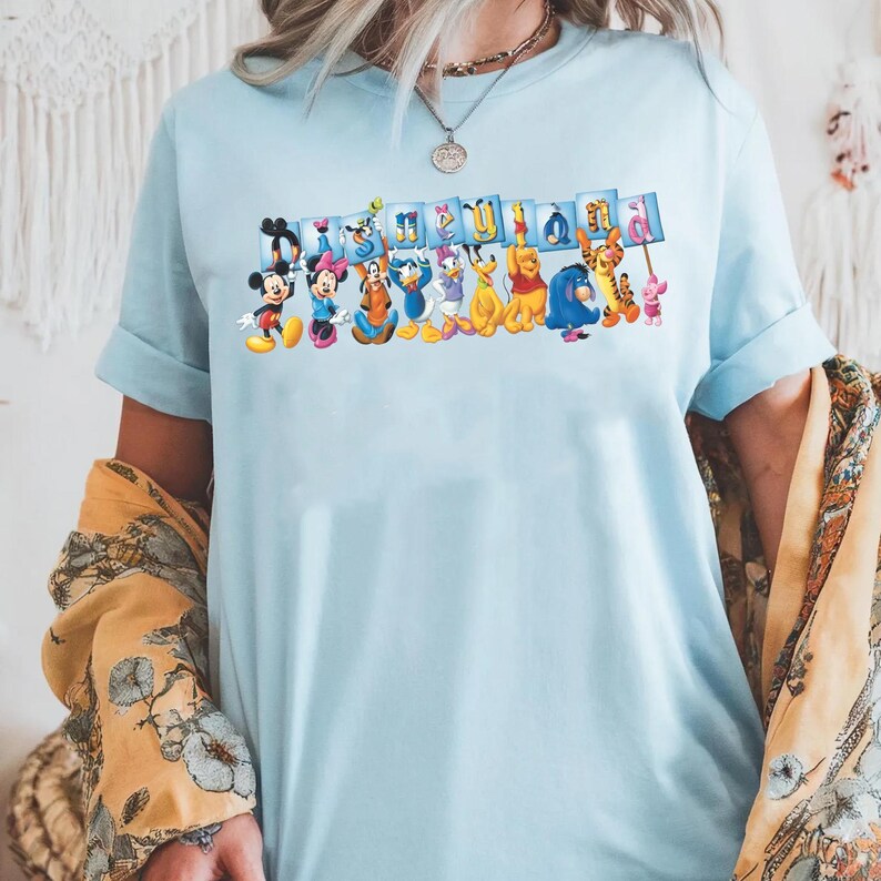 Puede incluir: Camiseta azul claro con un estampado colorido de personajes de Disney sosteniendo carteles que deletrean "It's a Small World". El dise&ntilde;o incluye a Mickey Mouse, Minnie Mouse, el Pato Donald y Winnie the Pooh. La camiseta tiene mangas cortas.