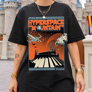 May include: Black t-shirt featuring a Star Wars Hyperspace Mountain graphic. The design includes the text "Star Wars" above "Hyperspace Mountain" in orange and blue. The graphic shows an X-wing fighter, a spaceship, and the Disneyland logo.