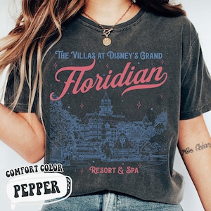 May include: Dark gray t-shirt with the text "The Villas at Disney's Grand Floridian Resort & Spa" in blue and pink. The shirt features a detailed blue illustration of the resort and the words "Comfort Color Pepper".