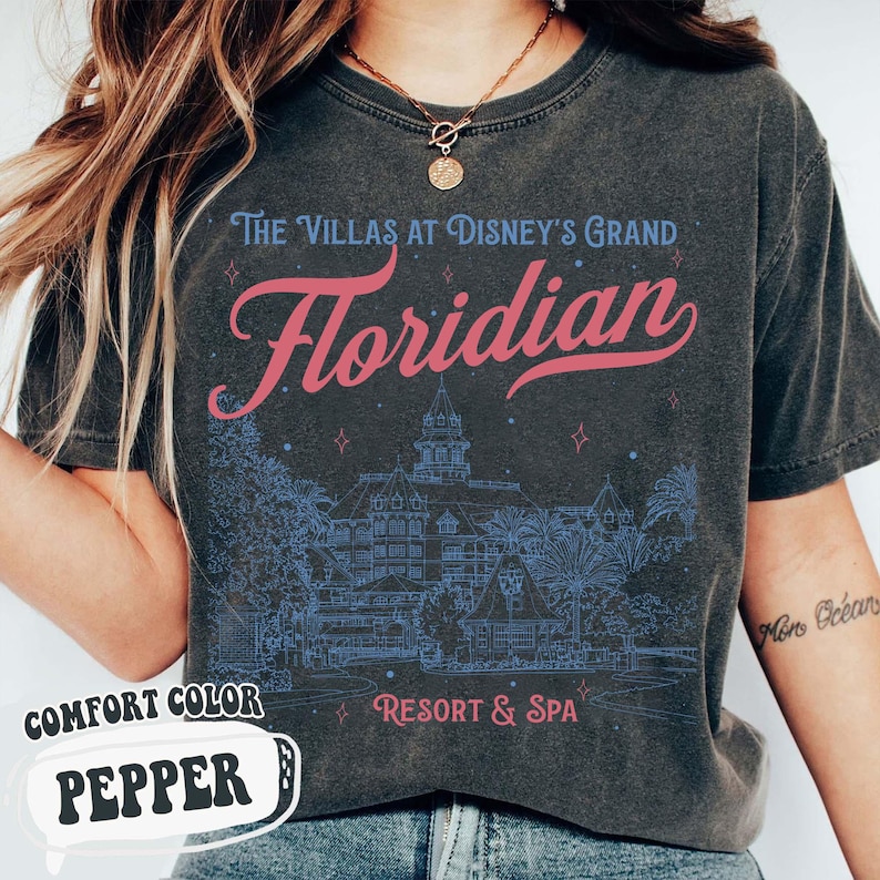 May include: Dark gray t-shirt with the text "The Villas at Disney's Grand Floridian Resort & Spa" in blue and pink. The shirt features a detailed blue illustration of the resort and the words "Comfort Color Pepper".