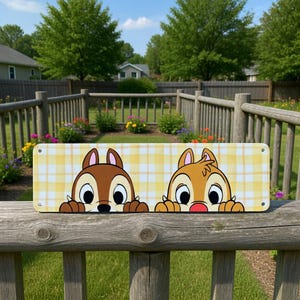 May include: A rectangular sign featuring Chip and Dale peeking over the edge. The background is a yellow and white plaid pattern. The sign is mounted on a wooden fence in an outdoor setting.