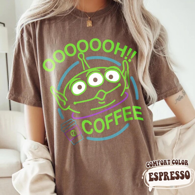 Puede incluir: Camiseta marr&oacute;n con un dise&ntilde;o de alien&iacute;gena verde y las palabras "OOOOH!! COFFEE". El alien&iacute;gena tiene tres ojos y est&aacute; rodeado por un c&iacute;rculo azul y morado. La camiseta est&aacute; etiquetada como "Comfort Color Espresso".