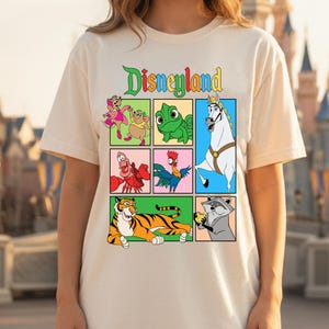Op de afbeelding: Crèmekleurig T-shirt met een kleurrijke afbeelding van Disneyland-personages in een rasterpatroon. Het woord "Disneyland" staat in groen, geel en oranje boven het raster. Het shirt heeft korte mouwen en een ronde hals.