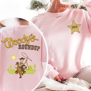 K&ouml;nnte beinhalten: Ein hellrosa Sweatshirt mit einem goldenen Sheriff-Abzeichen und dem Wort "Sheriff". Auf der R&uuml;ckseite des Sweatshirts stehen die Worte "Woody's Roundup" in Gelb und Braun sowie ein Cartoon-Cowboy auf einem Pferd.