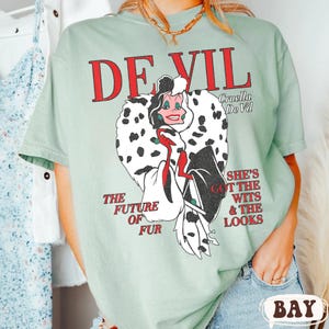 May include: A light green t-shirt featuring a graphic of Cruella De Vil with the text "DEVIL" in red. The graphic includes the text "THE FUTURE OF FUR" and "SHE'S GOT THE WITS & THE LOOKS".