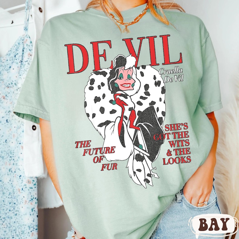 May include: A light green t-shirt featuring a graphic of Cruella De Vil with the text "DEVIL" in red. The graphic includes the text "THE FUTURE OF FUR" and "SHE'S GOT THE WITS & THE LOOKS".