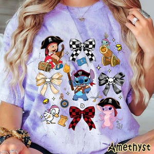 Może przedstawiać: Jasnofioletowa koszulka z postaciami pirat&oacute;w z kresk&oacute;wek i wzorami kokard. Koszulka przedstawia dziewczynę piratkę grającą na gitarze, Stitcha jako pirata i kaczkę w pirackim kapeluszu. Słowo "Amethyst" jest wydrukowane na dole.