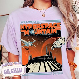 May include: Orchid-colored t-shirt featuring a retro-style graphic for Star Wars: Hyperspace Mountain at Disneyland. The design includes an X-wing fighter, the Millennium Falcon, and the Death Star against an orange and black background.