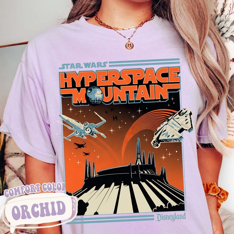 May include: Orchid-colored t-shirt featuring a retro-style graphic for Star Wars: Hyperspace Mountain at Disneyland. The design includes an X-wing fighter, the Millennium Falcon, and the Death Star against an orange and black background.