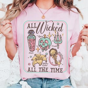 May include: Pink t-shirt with the text "ALL Wicked ALL THE TIME" in a rectangular design. The design includes illustrations of a potion bottle, a skull, a trident, a flower, and a golden orb, with character names.