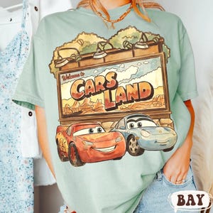 Op de afbeelding: Lichtgroen t-shirt met een afbeelding van Cars Land, met Lightning McQueen en Sally Carrera. Het bord leest "Welcome to Cars Land" in een woestijnlandschap. Het t-shirt is lichtgroen van kleur.