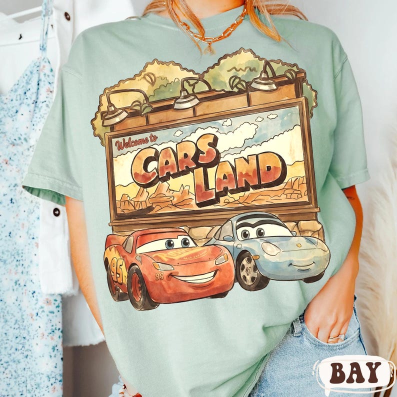 Op de afbeelding: Lichtgroen t-shirt met een afbeelding van Cars Land, met Lightning McQueen en Sally Carrera. Het bord leest "Welcome to Cars Land" in een woestijnlandschap. Het t-shirt is lichtgroen van kleur.