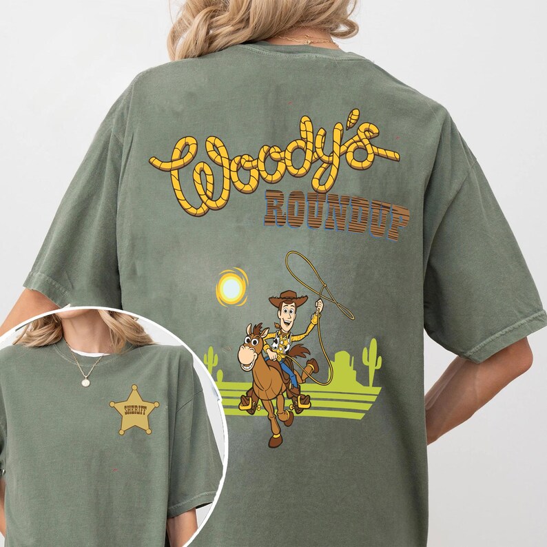 K&ouml;nnte beinhalten: Olivgr&uuml;nes T-Shirt mit einer Cartoon-Illustration von Woody, der mit einem Lasso auf einem Pferd reitet, einer Sonne und Kakteen. Der Text "Woody's Roundup" ist in Gelb und Braun auf der R&uuml;ckseite aufgedruckt.