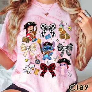 Może przedstawiać: R&oacute;żowa koszulka typu tie-dye z r&oacute;żnymi postaciami pirat&oacute;w z kresk&oacute;wek i wzorami kokard. Koszulka zawiera obrazy pirata Stitcha, kaczki i innych ilustracji o tematyce pirackiej. Słowo "Clay" jest wydrukowane w prawym dolnym rogu.