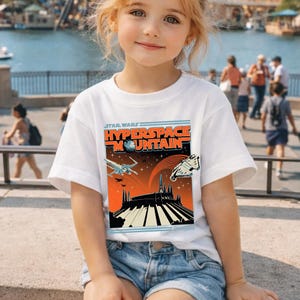 May include: White t-shirt featuring a Star Wars Hyperspace Mountain graphic. The design includes spaceships, a mountain silhouette, and the Disneyland logo. The shirt is worn by a young person.