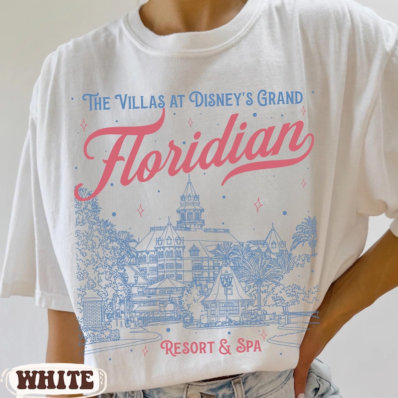 May include: White t-shirt with a graphic design featuring the text "The Villas at Disney's Grand Floridian Resort & Spa" in blue and pink. The design includes an illustration of a resort building and palm trees.
