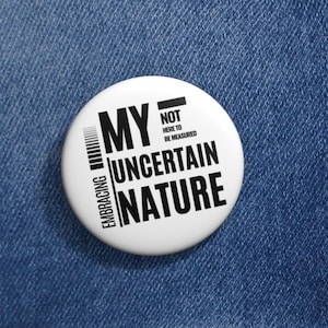 May include: A white button pin with black text that reads "MY UNCERTAIN NATURE" and "EMBRACING" vertically along the left side. The text "NOT HERE TO BE MEASURED" is also on the pin. The pin is on a blue denim jacket.