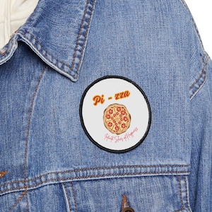 May include: A woman wearing a blue denim jacket with a white hoodie underneath. She is wearing a round, white patch with a red and orange pizza graphic and the text "Pi-zza" in a retro font. The text below the pizza says "Hot Slices of Happiness".