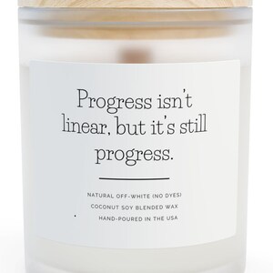 May include: A white candle in a frosted glass jar with a wooden lid. The label reads "Progress isn't linear, but it's still progress." The candle is made with natural off-white wax and hand-poured in the USA.