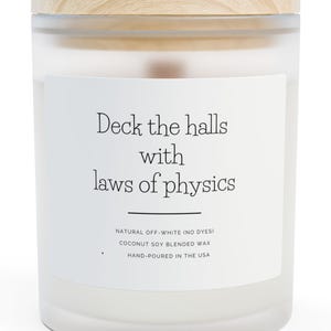 May include: A white candle in a glass jar with a wooden lid. The label reads "Deck the halls with laws of physics." Additional text states the candle is natural, off-white, and hand-poured in the USA.