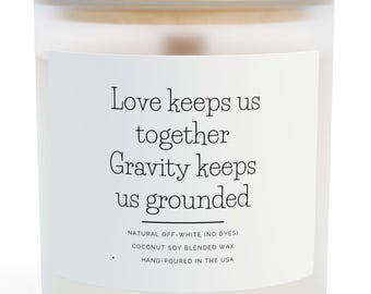 Puede incluir: Una vela blanca en un tarro de vidrio esmerilado con tapa de madera. La etiqueta dice "Love keeps us together, Gravity keeps us grounded". La vela está hecha con cera de soja de coco y vertida a mano en los EE. UU.